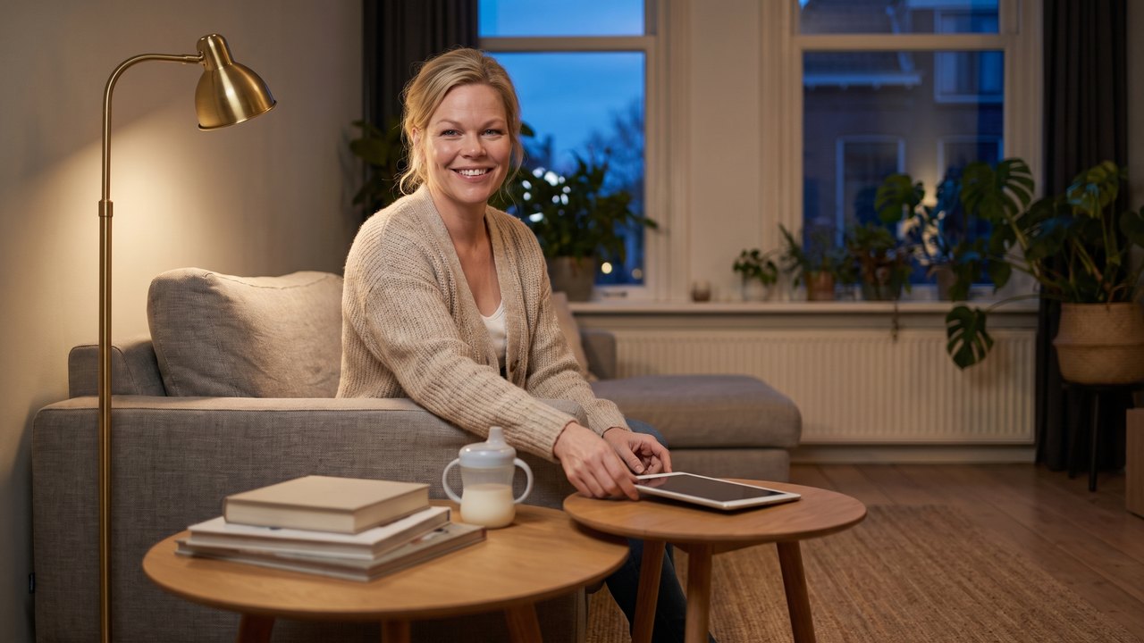 Evening in a Dutch terraced house: a fair haired woman in a knitted cardigan sits on a grey sofa and smiles at the camera, her hand resting on a dark tablet lying on a small round oak side table; on a second side table stand a toddler's cup of milk and a stack of books, with a brass floor lamp beside her and tall windows full of blue dusk behind.