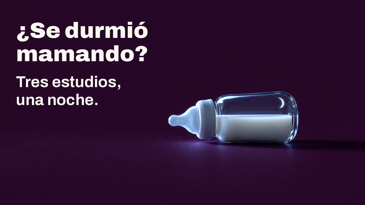 ¿Se durmió mamando? Tres estudios, una noche. Un pequeño biberón de cristal con leche yace de lado sobre un fondo ciruela profundo, iluminado desde un lado por la luz fría de una pantalla que no se ve, con una fina luna creciente reflejada en el cristal.
