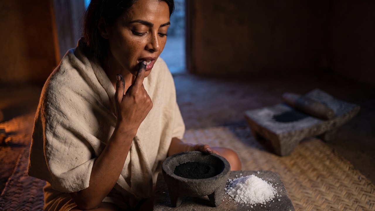 Is charcoal toothpaste good for your teeth: a Mexica woman in a house in Tenochtitlan presses a charcoal blackened fingertip along her own gum, a three legged basalt bowl of black charcoal dust and a heap of white salt beside her