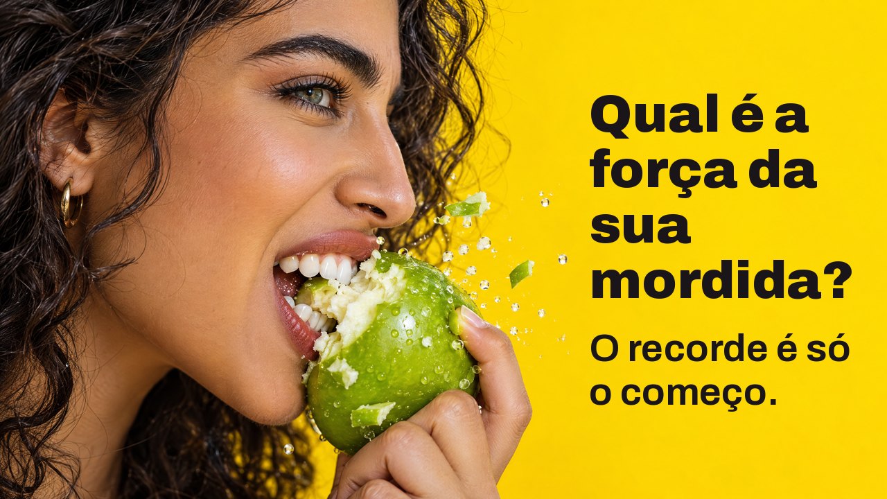 Qual é a força da sua mordida? O recorde é só o começo. Grande plano de perfil de uma mulher a morder com força uma maçã verde estaladiça, com pedaços a voar, sobre amarelo vivo.