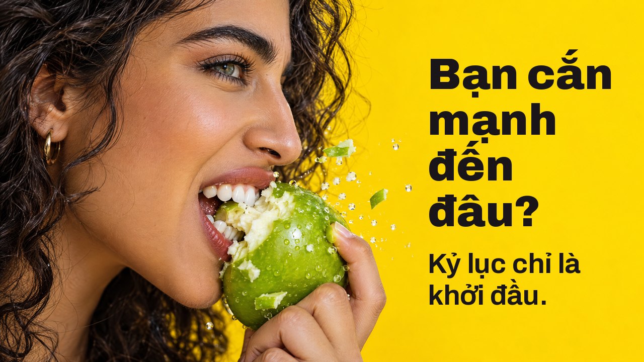 Bạn cắn mạnh đến đâu? Kỷ lục chỉ là khởi đầu. Cận cảnh nghiêng một người phụ nữ cắn mạnh quả táo xanh giòn, mảnh táo văng ra, trên nền vàng rực.